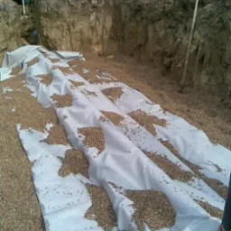 Wykop pod oczyszczalnię ścieków IN-DRAN z ułożoną geowłókniną i zasypywany żwirem, widoczny pracownik z łopatą w tle.