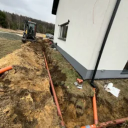 Pomarańczowe rury kanalizacyjne ułożone w wykopie obok budynku jednorodzinnego; minikoparka w tle, widoczne narzędzia i rolka papieru.