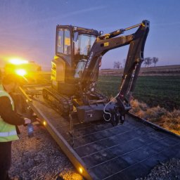 PW LARIS ANNA MOŁCZAN - Mała koparka Volvo na platformie transportowej z włączonymi światłami ostrzegawczymi, operator w kamizelce odblaskowej, w tle pole i zachodzące słońce.