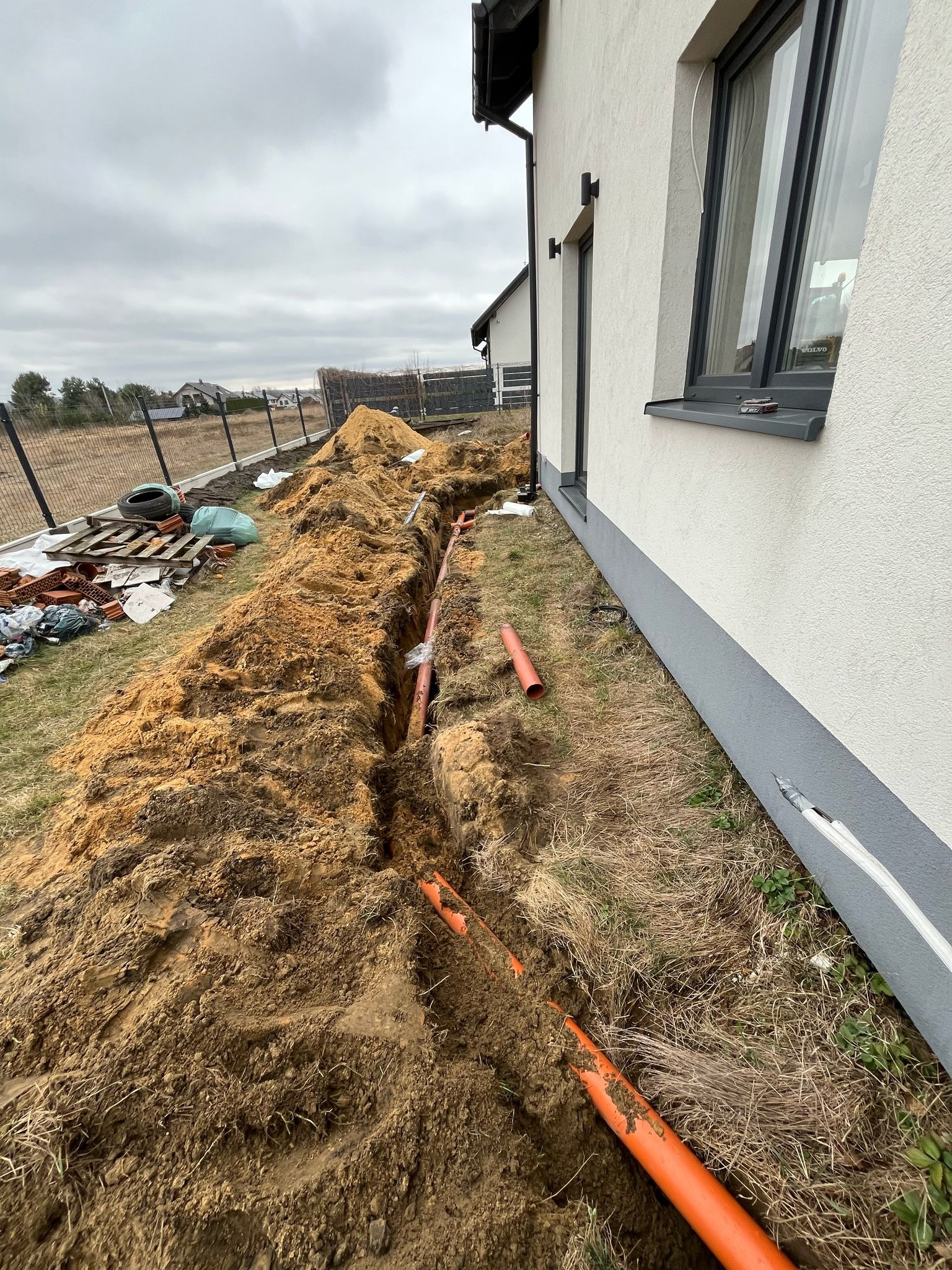 Wykop pod instalacje biegnący wzdłuż ściany budynku, z widocznymi pomarańczowymi rurami, ziemią i fragmentem ogrodzenia w tle, pochmurne niebo.