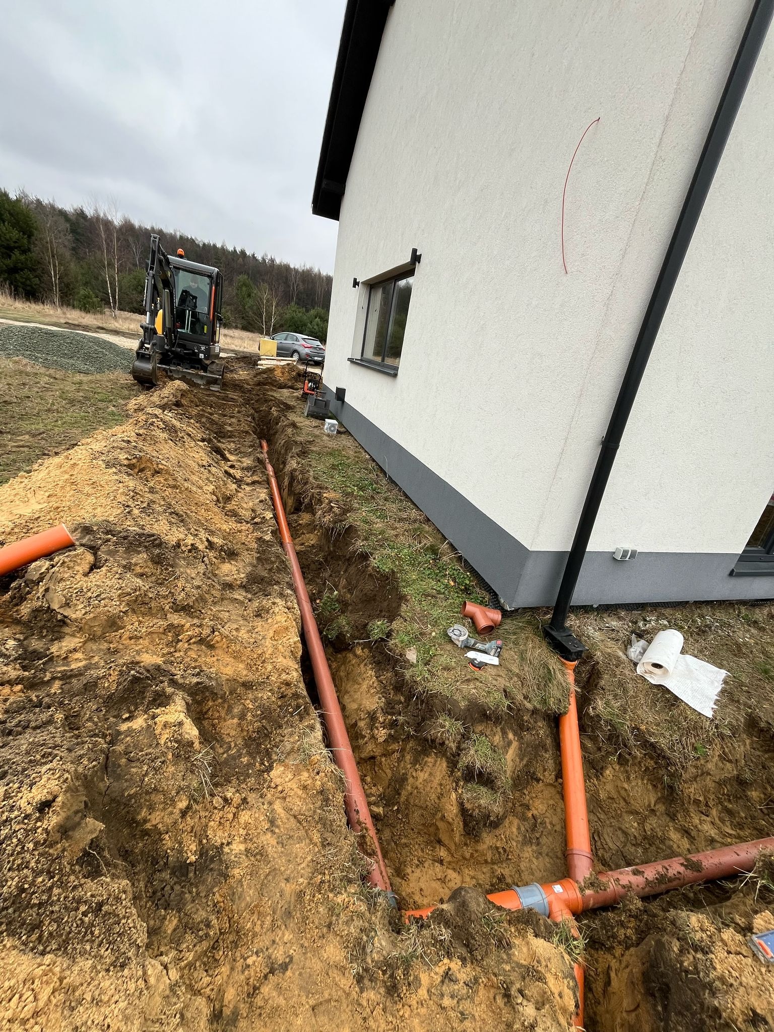 Pomarańczowe rury kanalizacyjne ułożone w wykopie obok budynku jednorodzinnego; minikoparka w tle, widoczne narzędzia i rolka papieru.
