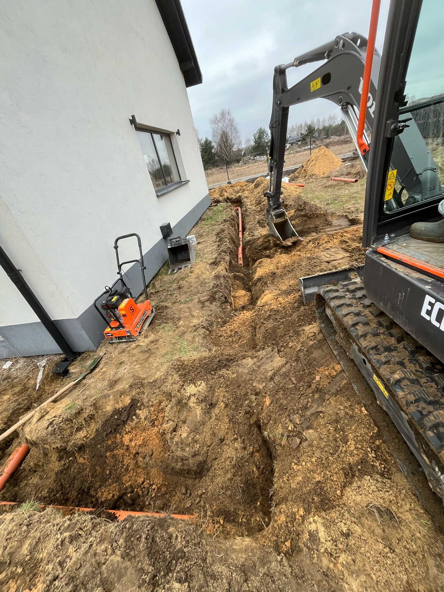 Wąski wykop pod rury w ziemi wzdłuż ściany budynku, z widocznym minikoparką i zagęszczarką w tle.