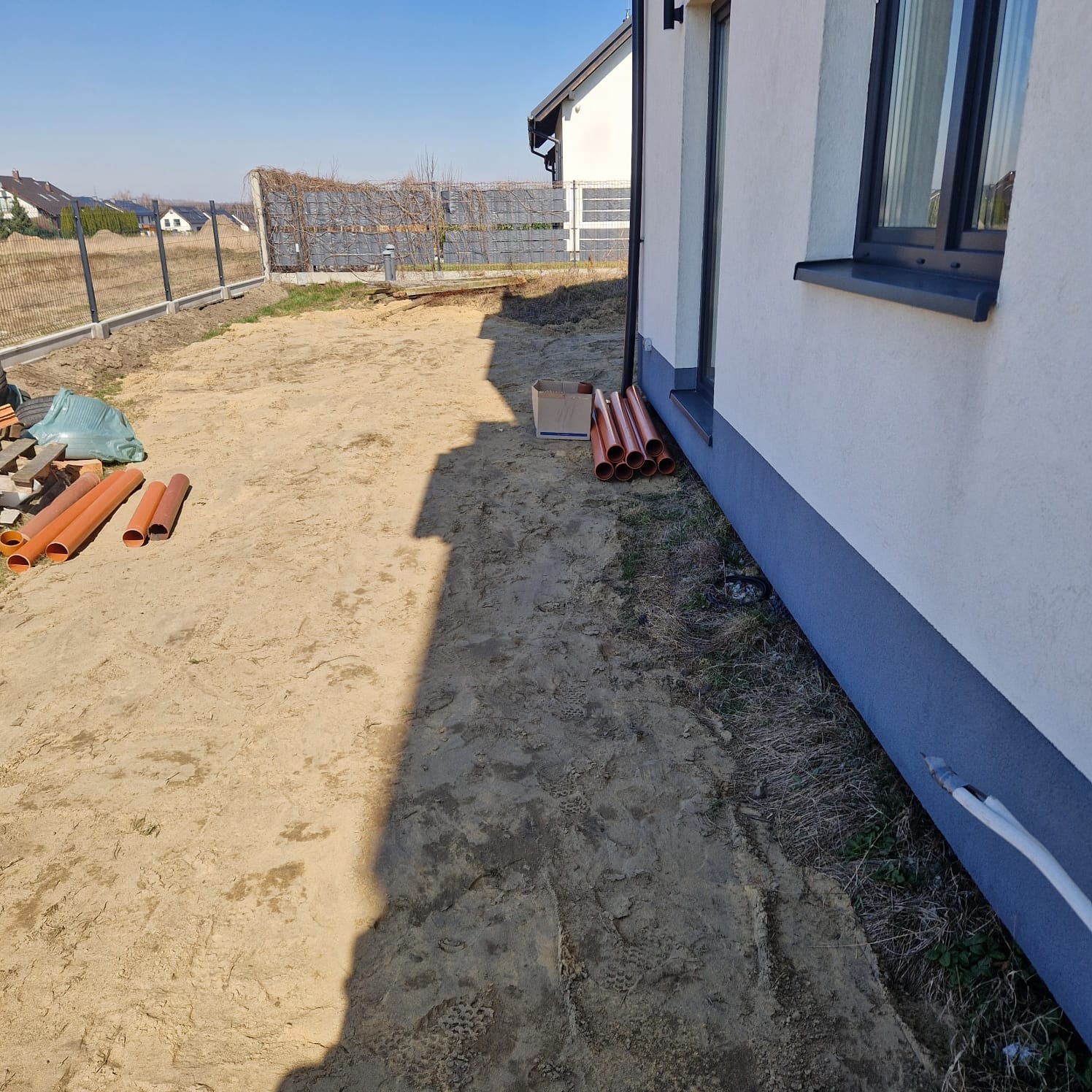 Przygotowany teren pod budowę z widocznymi rurami drenarskimi, piaskiem i częściowo wykończonym budynkiem w tle, oświetlony jasnym światłem słonecznym, ukazujący etap prac ziemnych.