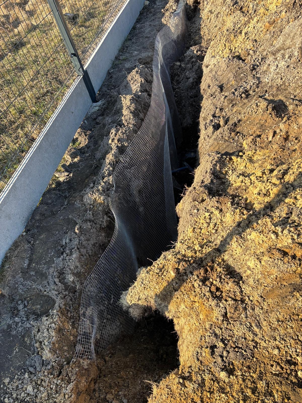 Wykop w ziemi zabezpieczony siatką metalową, widoczny fragment ogrodzenia z betonową podmurówką.