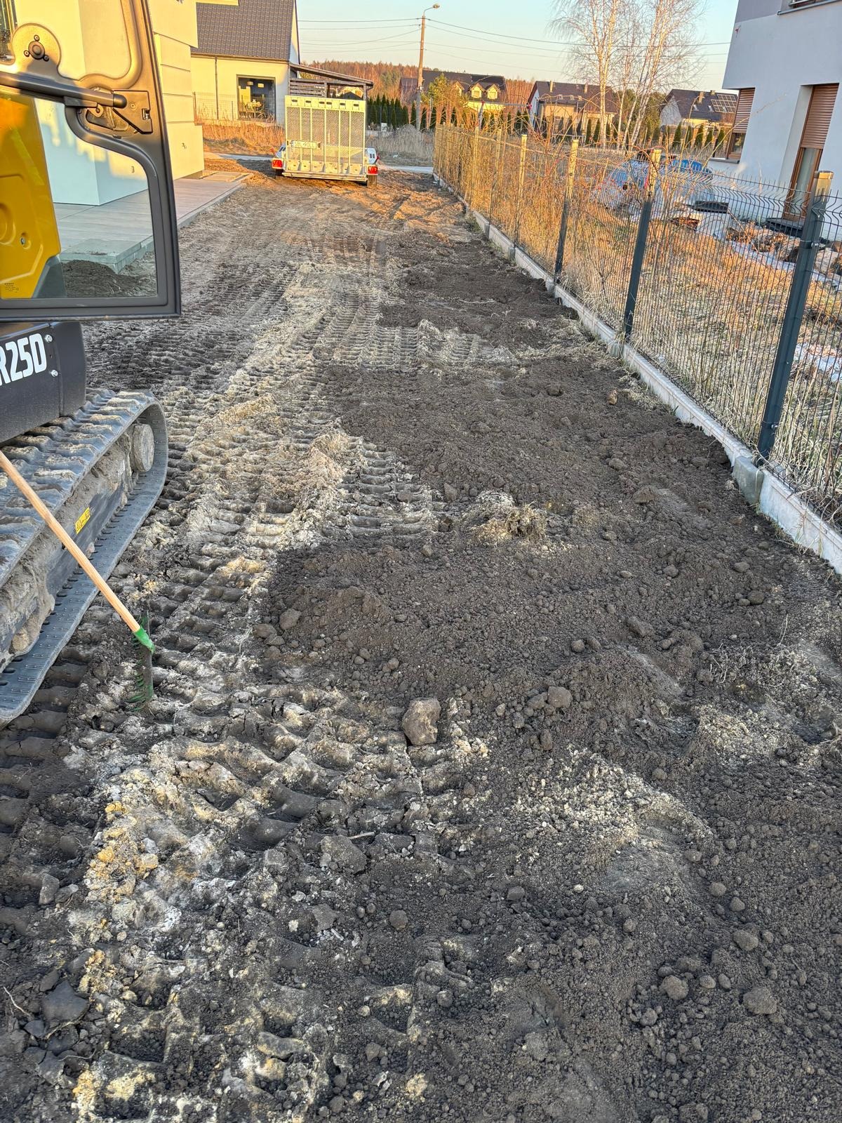 Świeżo wyrównany teren pod budowę z widocznymi śladami gąsienic minikoparki i fragmentem ogrodzenia panelowego w tle.