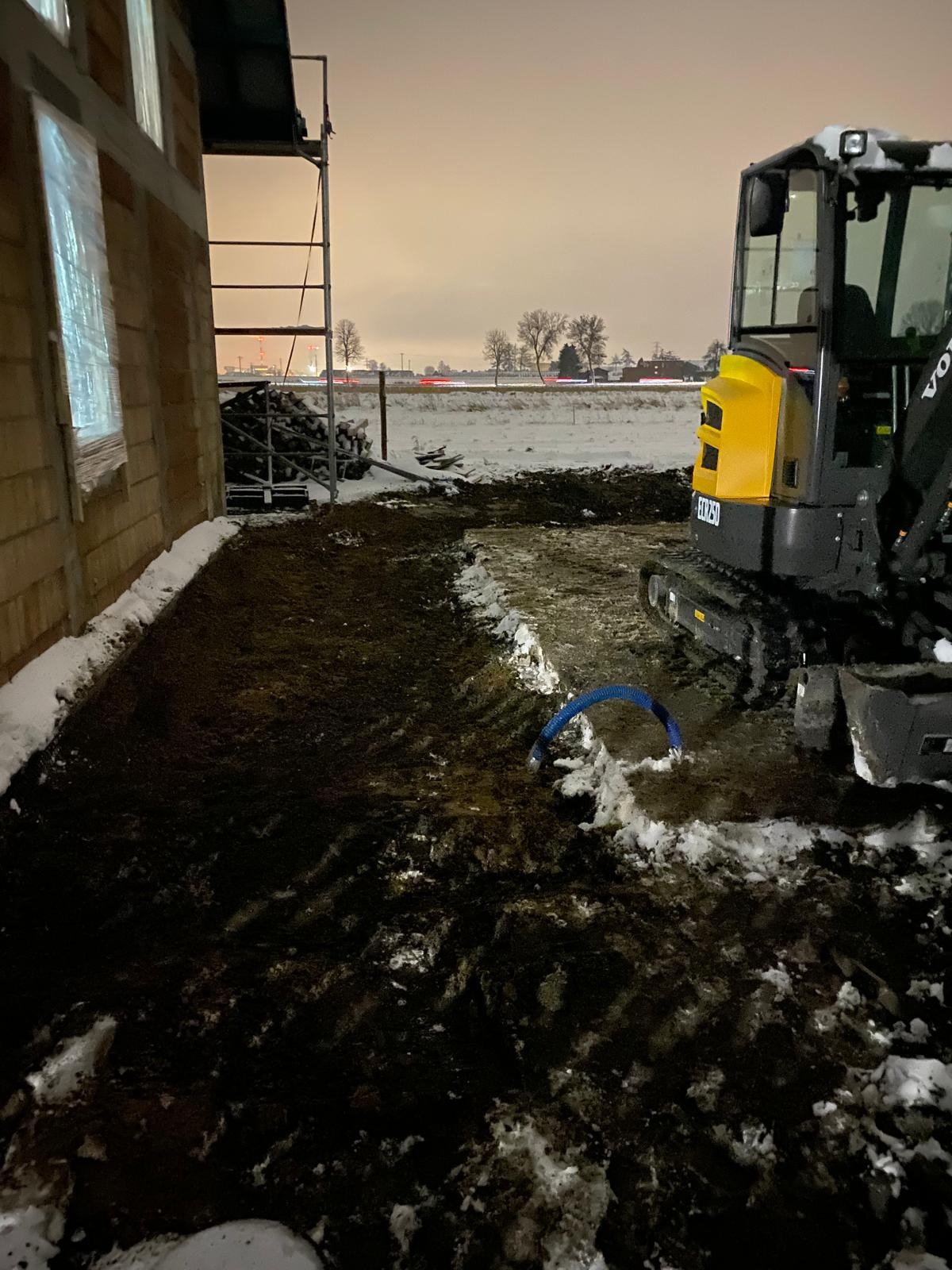Wykop pod instalacje w zimowej scenerii, widoczny fragment budynku w stanie surowym, ułożona rura instalacyjna oraz zaparkowana koparka Volvo EC25D, oświetlenie nocne.