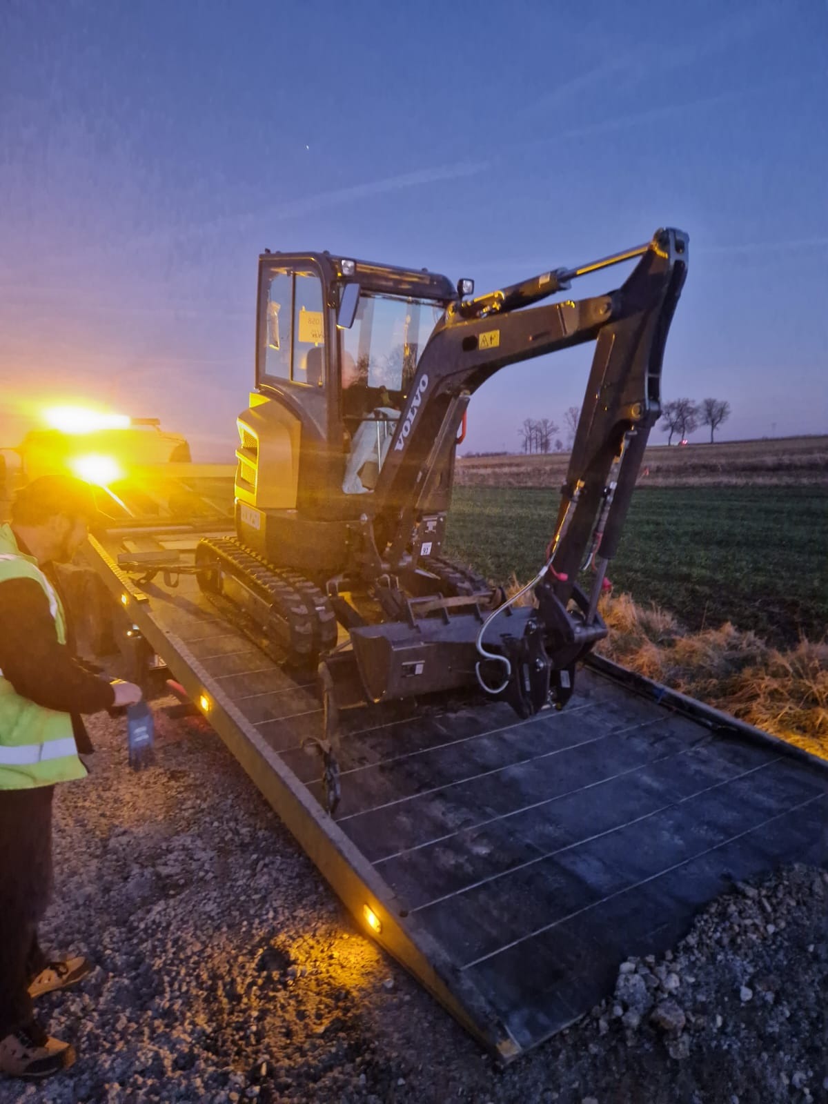 Mała koparka Volvo na platformie transportowej z włączonymi światłami ostrzegawczymi, operator w kamizelce odblaskowej, w tle pole i zachodzące słońce.