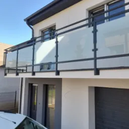 Nowoczesny balkon z czarną metalową ramą i mlecznym szkłem, widok z dołu na tle jasnej fasady domu z grafitowymi akcentami.