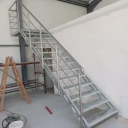 Metalowe schody z nierdzewną balustradą w hali przemysłowej, widoczne rusztowanie i narzędzia malarskie.