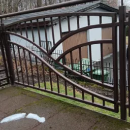 Brązowa brama wjazdowa z ozdobnymi elementami na tle garażu i drzew bez liści, z fragmentem topniejącego śniegu na brukowanej powierzchni.