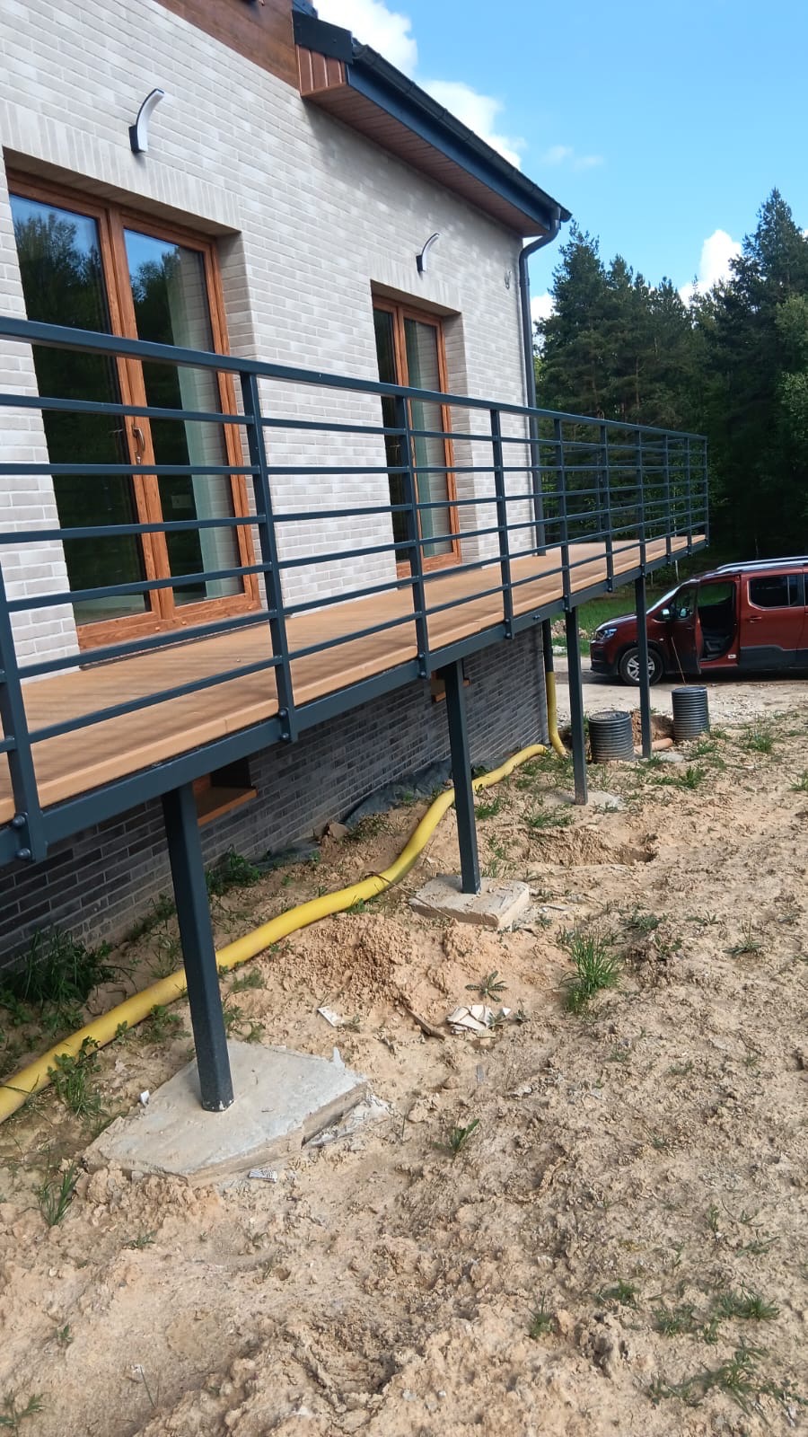 Nowoczesna balustrada z poziomych prętów, zamontowana na drewnianym tarasie przy domu z jasnej cegły, widoczna z zewnątrz w trakcie prac wykończeniowych.