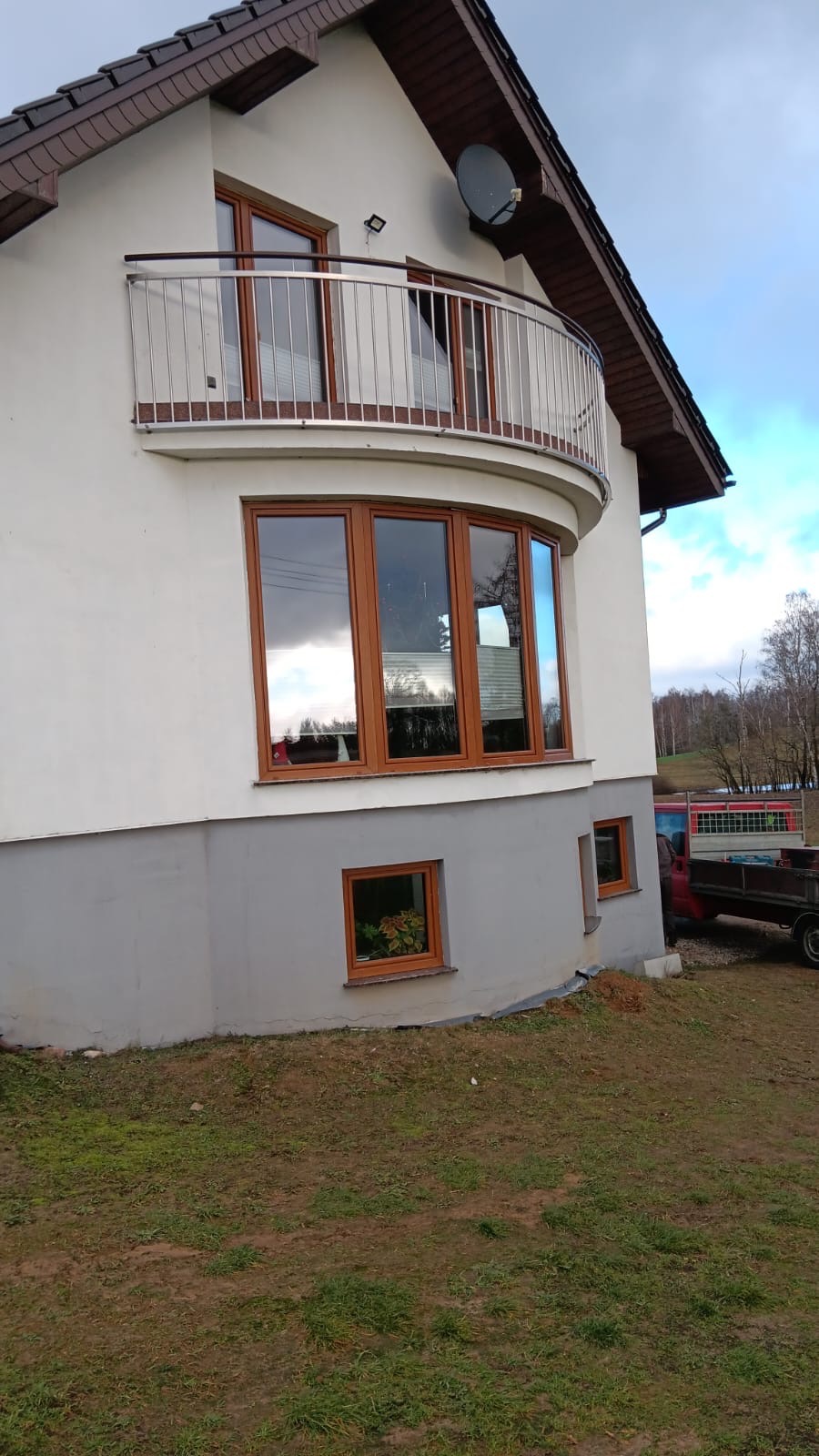 Biały dom z brązowymi oknami i balkonem z nierdzewną balustradą, widok z boku na tle pochmurnego nieba i drzew