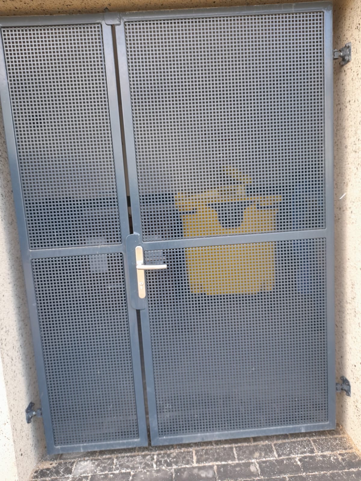 Szara, metalowa brama wjazdowa z perforowanymi panelami i srebrną klamką, widziana z bliska, z widocznym żółtym elementem w tle.