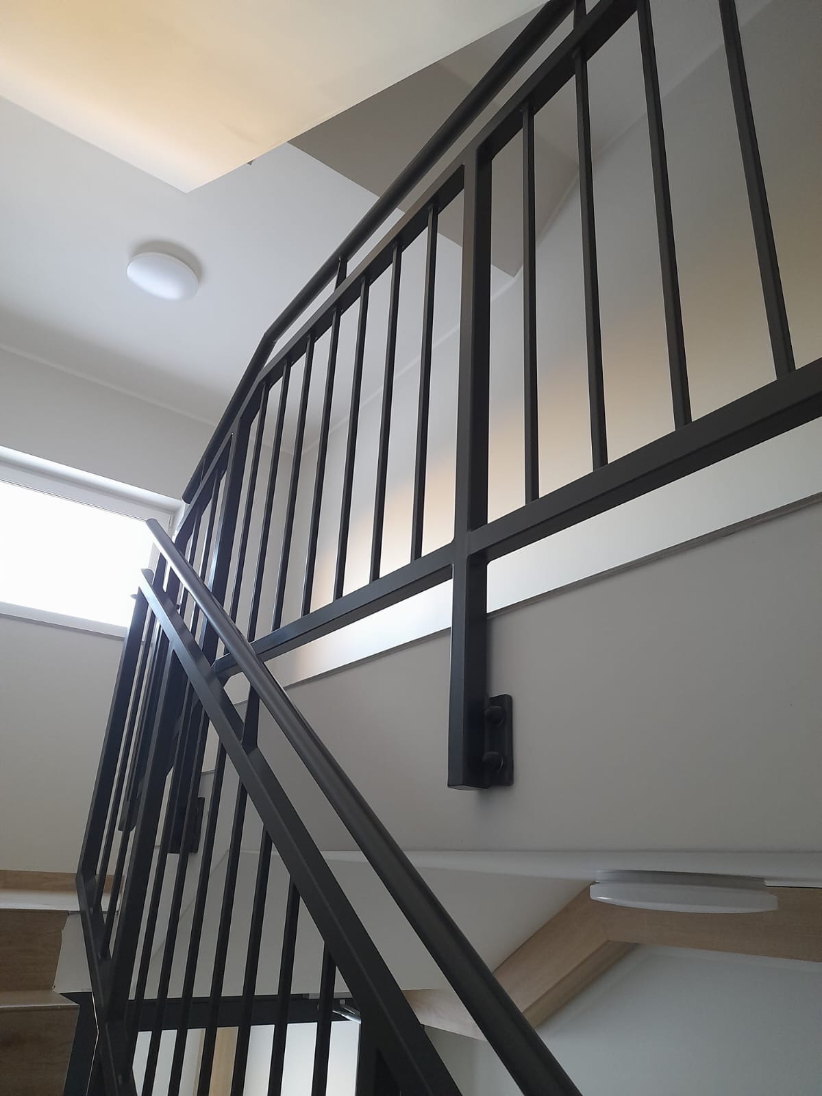 Nowoczesna, ciemnoszara balustrada schodowa z pionowymi szczeblami, mocowana do ściany za pomocą czarnych, metalowych uchwytów, widoczna z dołu, przy oświetleniu sufitowym.