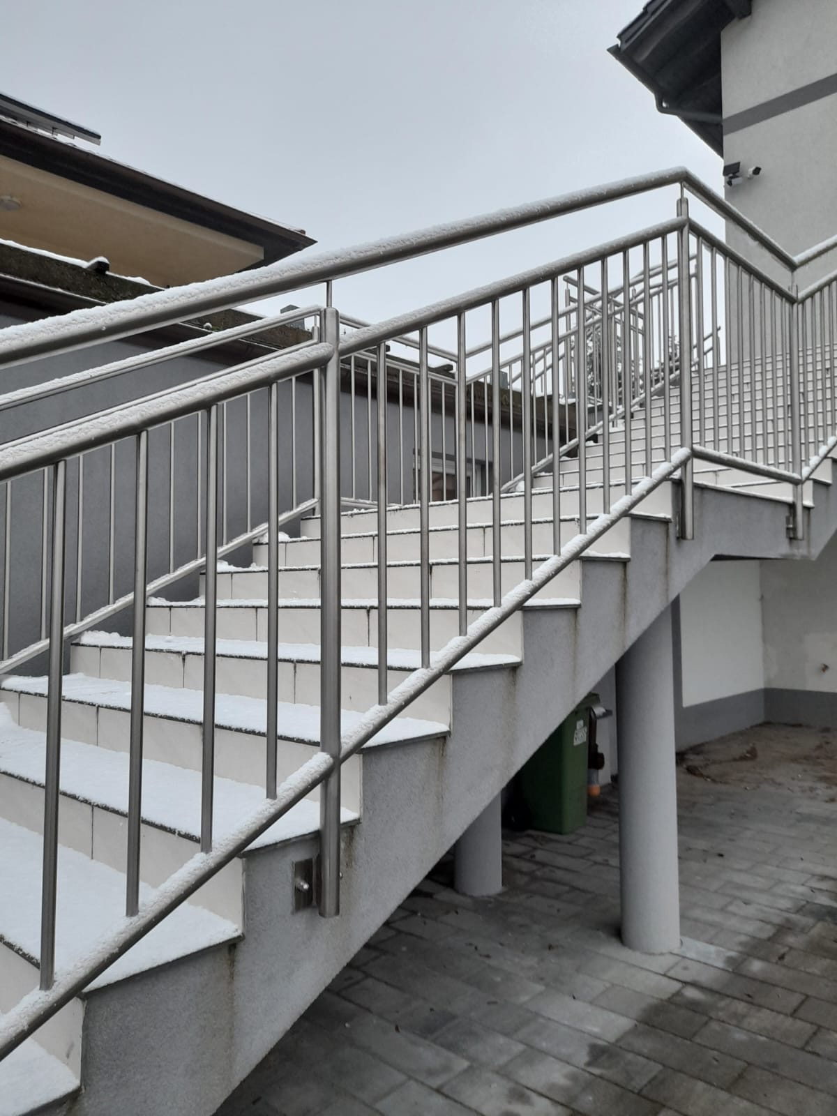 Zewnętrzne schody z białymi stopniami i stalową balustradą, pokryte cienką warstwą śniegu, prowadzące na wyższą kondygnację budynku.