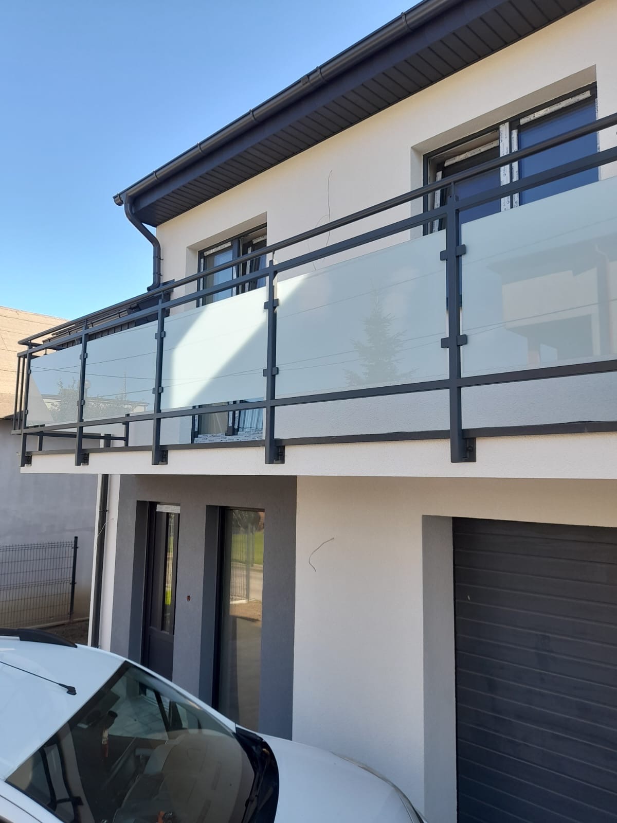 Nowoczesny balkon z czarną metalową ramą i mlecznym szkłem, widok z dołu na tle jasnej fasady domu z grafitowymi akcentami.