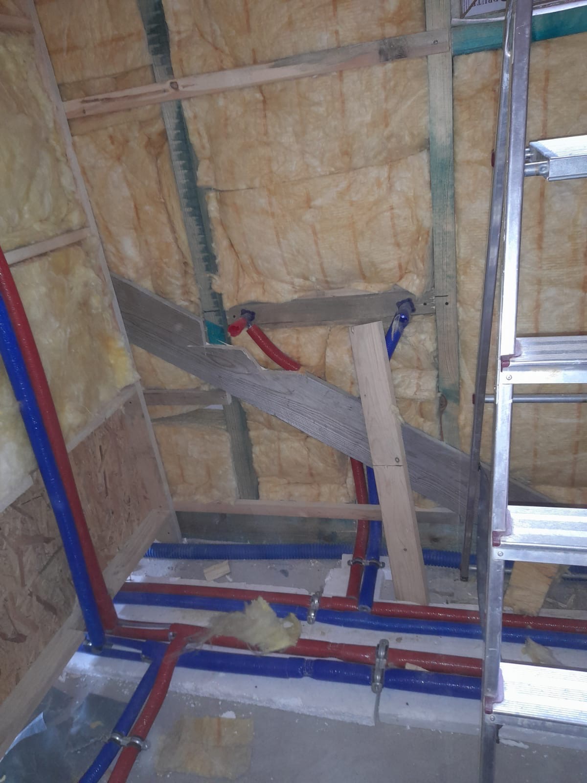 Montaż instalacji wodnej w ocieplonym poddaszu, widoczne rury PEX w kolorze niebieskim i czerwonym, mocowane do drewnianej konstrukcji dachu, obok drabina aluminiowa.