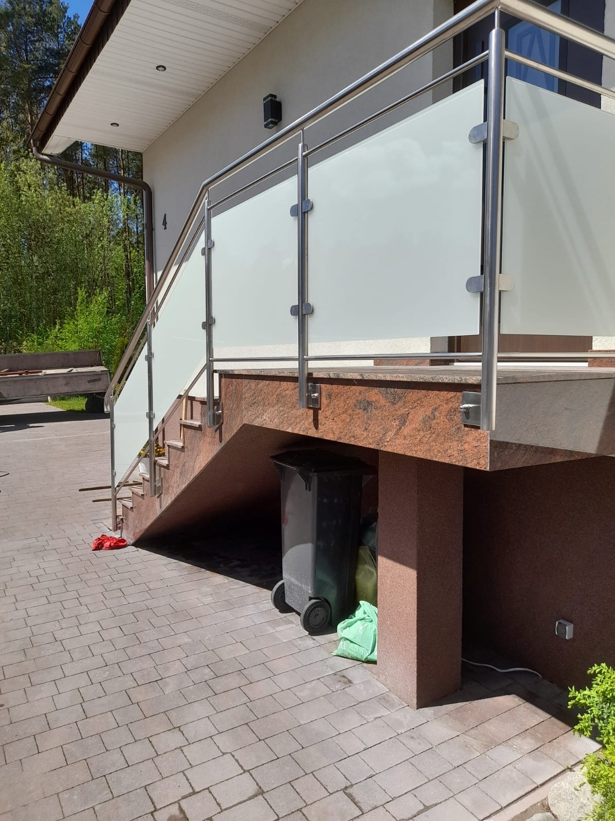 Zewnętrzne schody z balustradą ze stali nierdzewnej i mlecznego szkła, widok na posesję z kostki brukowej i kosz na śmieci pod schodami.