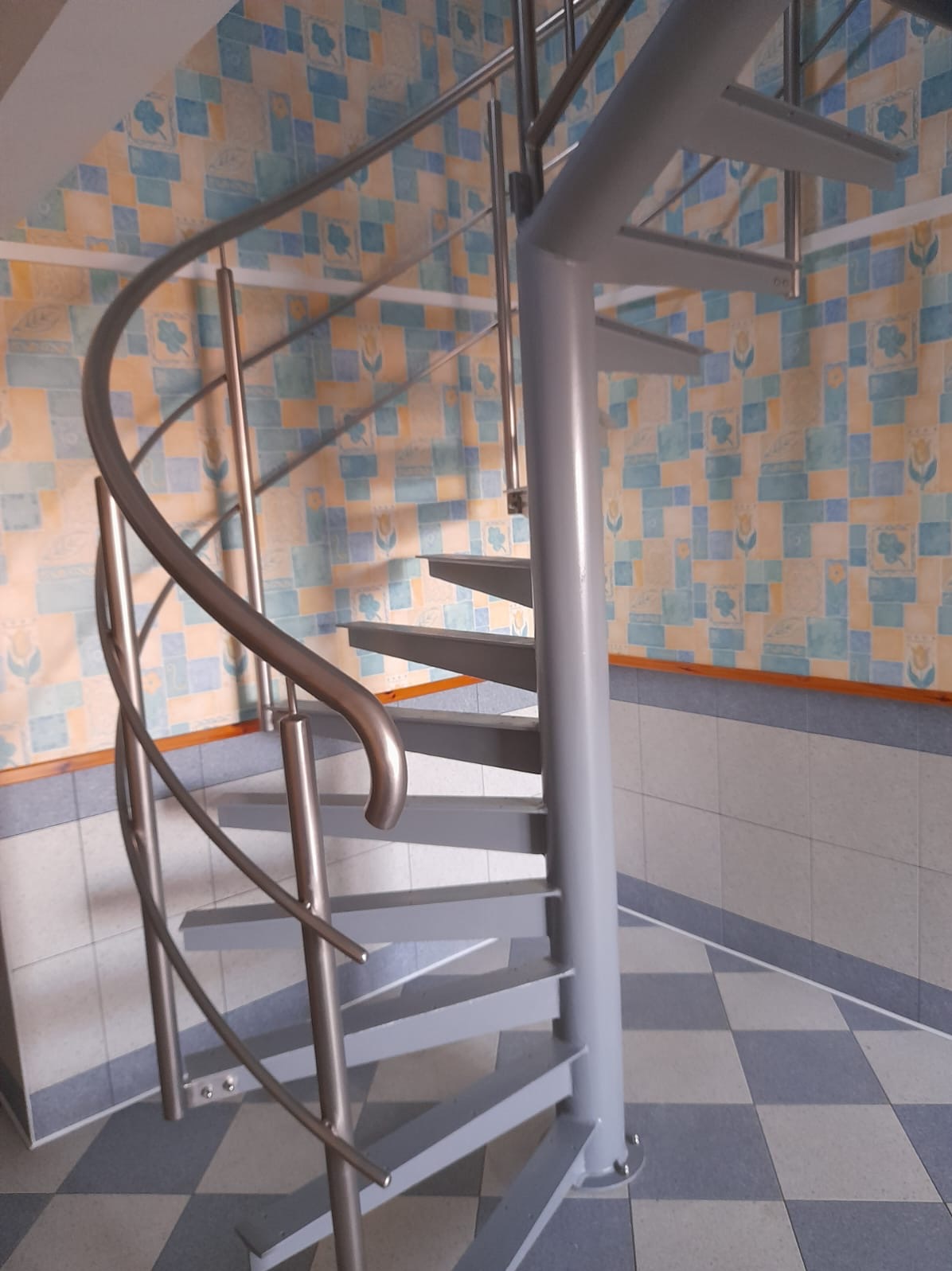 Metalowe schody spiralne wewnątrz budynku, z szarą konstrukcją i nierdzewną balustradą, na tle ściany wyłożonej wzorzystymi płytkami.