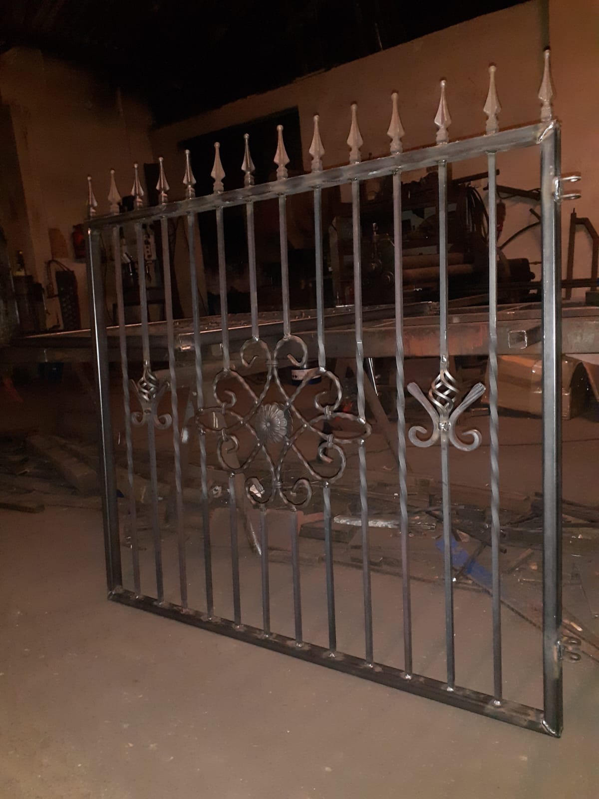 Surowa, metalowa konstrukcja bramy z dekoracyjnymi elementami kwiatowymi i ostrzami na górze, widoczna w warsztacie na tle maszyn.