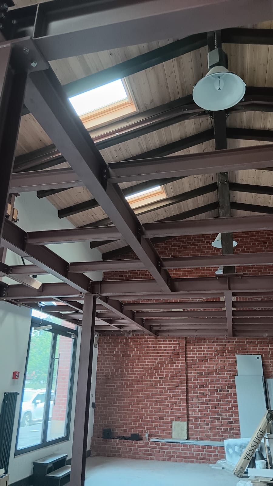 Wnętrze budynku w trakcie budowy z widoczną stalową konstrukcją nośną, drewnianym stropem z oknami dachowymi oraz fragmentem ściany z czerwonej cegły.