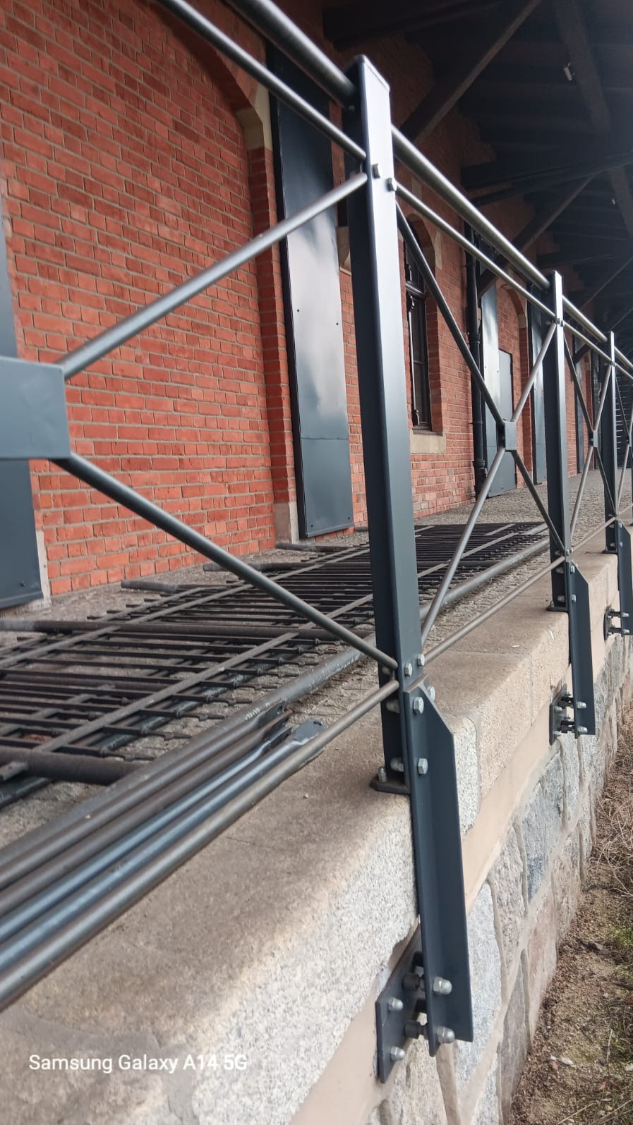 Metalowa balustrada w kolorze grafitowym, zamocowana na kamiennym murze, z widoczną ceglaną ścianą budynku w tle.