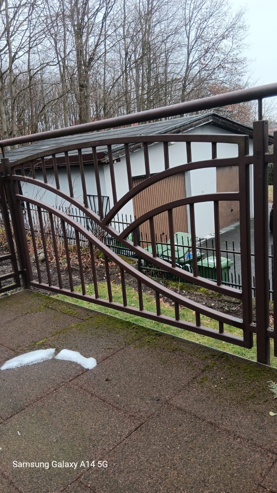 Brązowa brama wjazdowa z ozdobnymi elementami na tle garażu i drzew bez liści, z fragmentem topniejącego śniegu na brukowanej powierzchni.