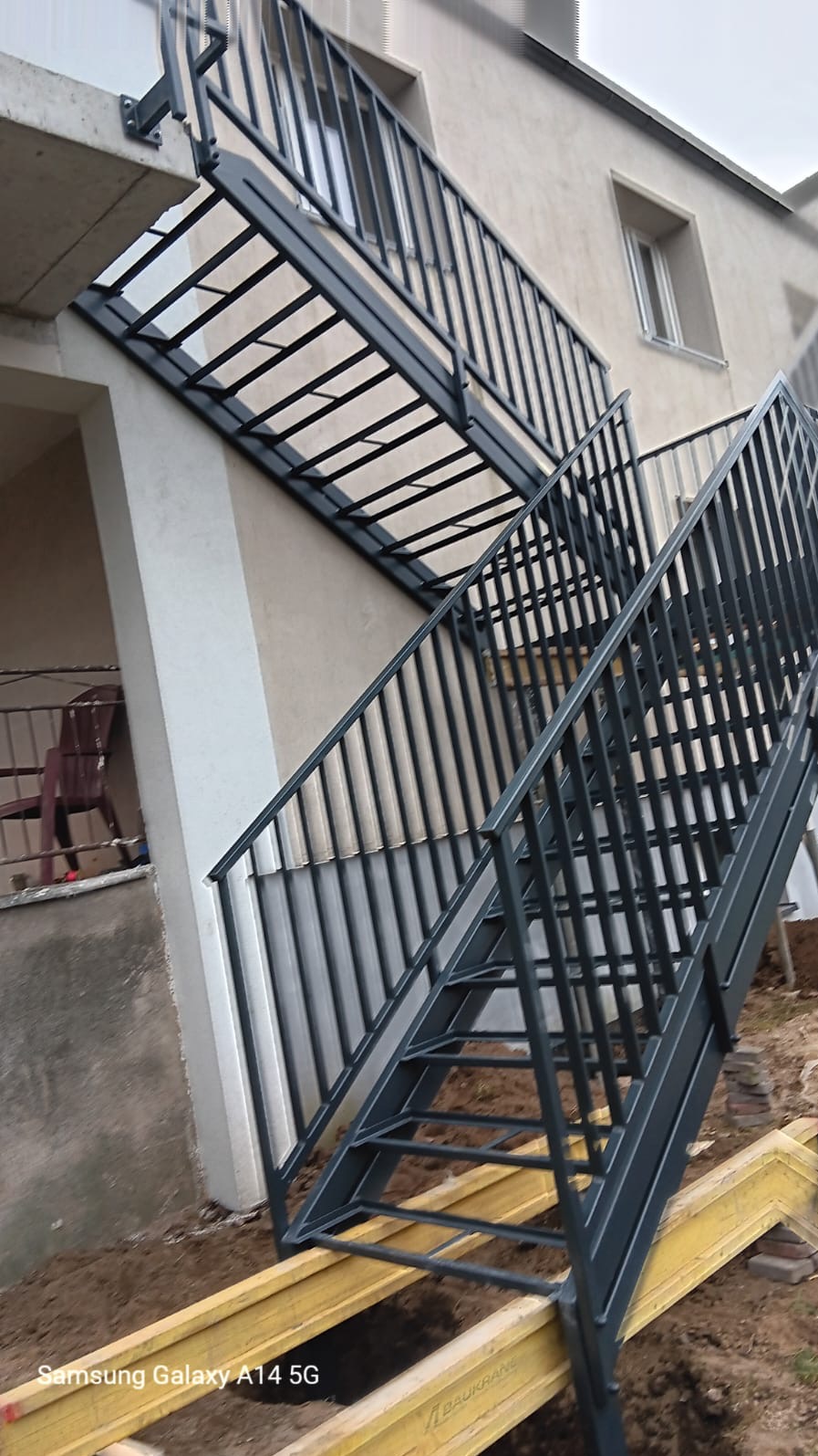 Zewnętrzne schody stalowe z czarną balustradą, prowadzące na balkon w budynku o beżowej elewacji, widoczne żółte elementy konstrukcyjne u podstawy schodów.