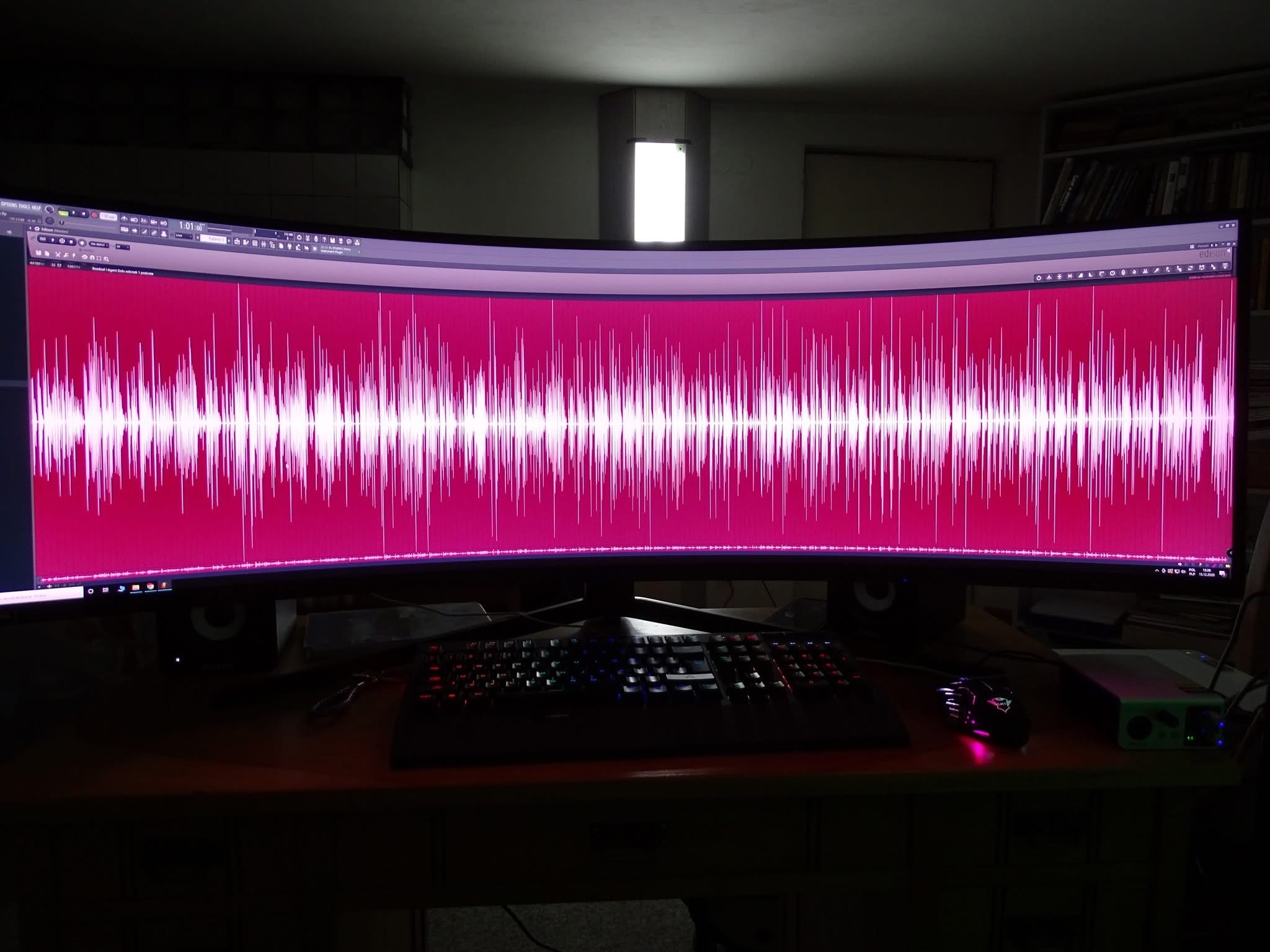 Ultraszeroki monitor wyświetla edytor audio z różowym waveform. Widoczna klawiatura z podświetleniem RGB, głośniki i interfejs audio na biurku.