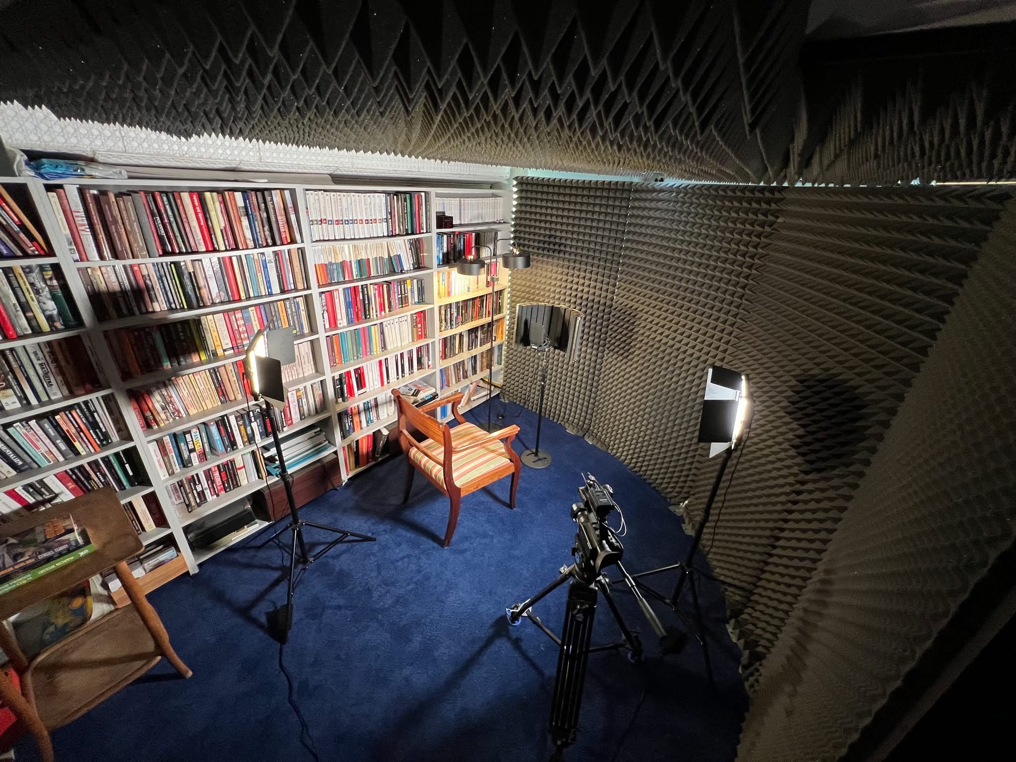 Studio nagrań w Warszawie: Pokój wygłuszony piramidkami, kamera na statywie, oświetlenie, fotel i regał z książkami tworzą profesjonalne tło.