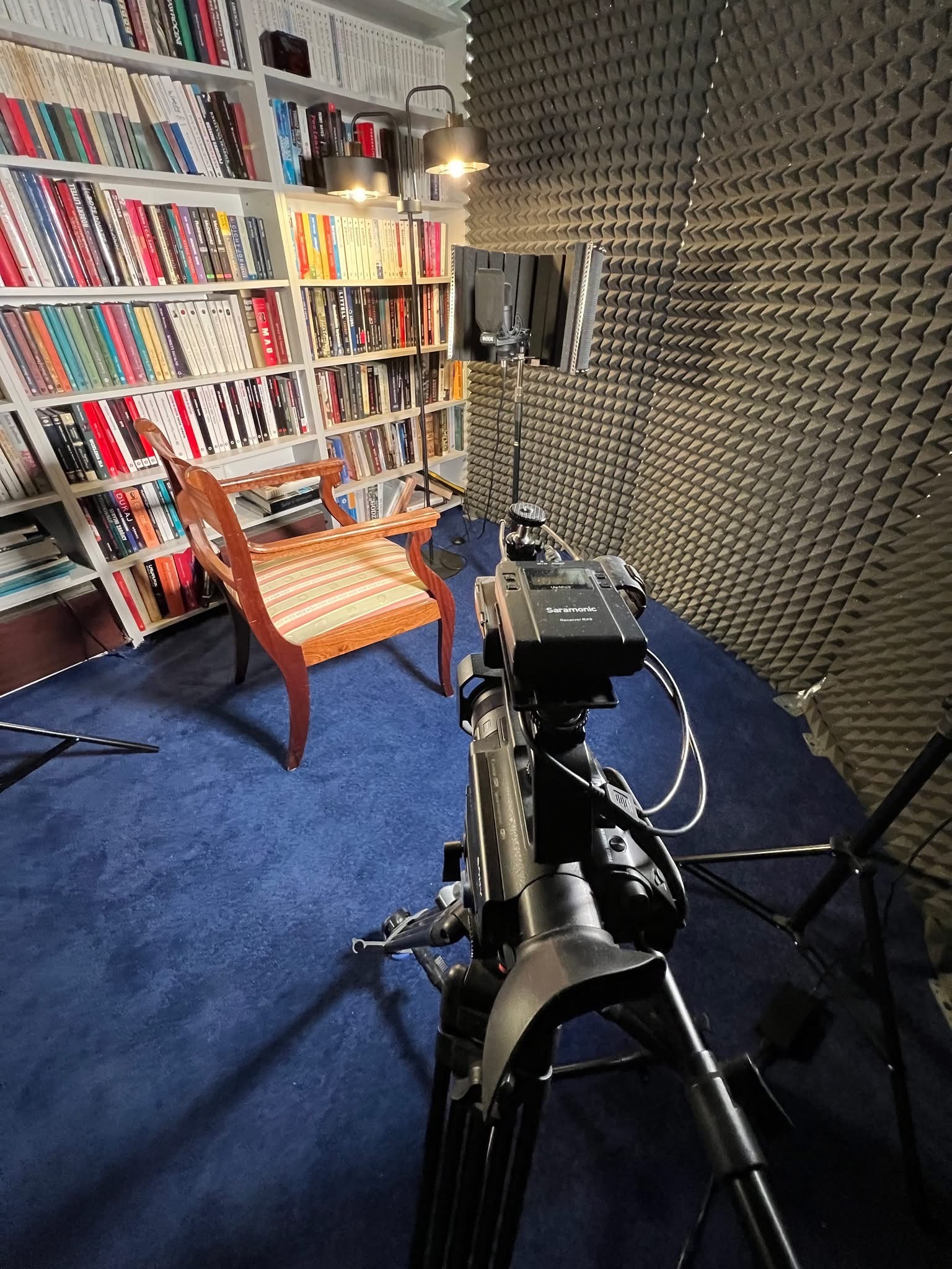 Kamera na statywie w domowym studiu nagrań z wygłuszonymi ścianami i regałem z książkami w tle. Przygotowanie do nagrania filmu.