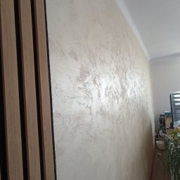 Marcin Polkowski - Ściana z dekoracyjną, beżową fakturą i pionowymi panelami drewnianymi z czarnymi wstawkami, widoczne z boku, z fragmentem rośliny w tle.