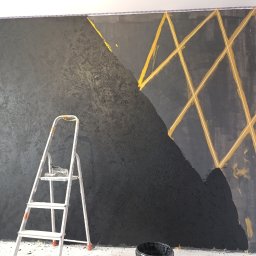 Marcin Polkowski - Ściana w trakcie malowania na czarno z geometrycznym wzorem z taśmy malarskiej, widoczna drabina malarska i wiadro z farbą.