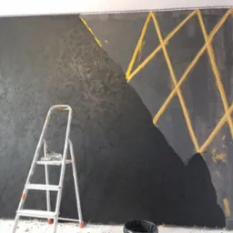 Ściana w trakcie malowania na czarno z geometrycznym wzorem z taśmy malarskiej, widoczna drabina malarska i wiadro z farbą.