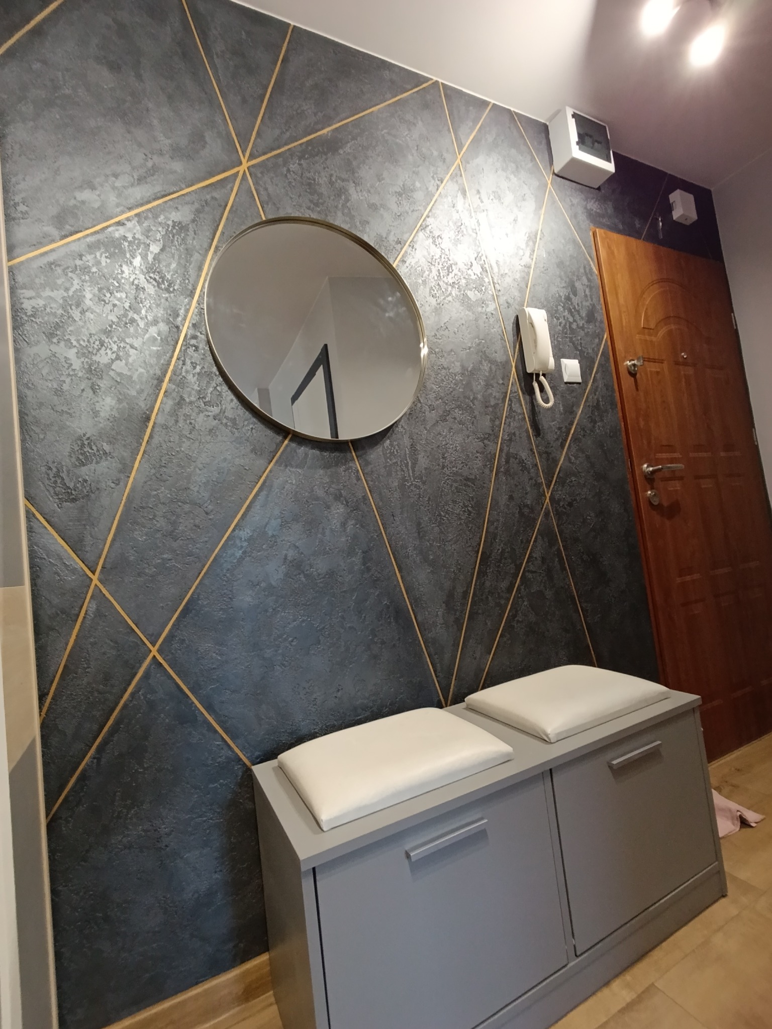 Szara ściana w przedpokoju z dekoracyjnym malowaniem w geometryczne złote linie, okrągłe lustro, szara szafka na buty z siedziskiem.