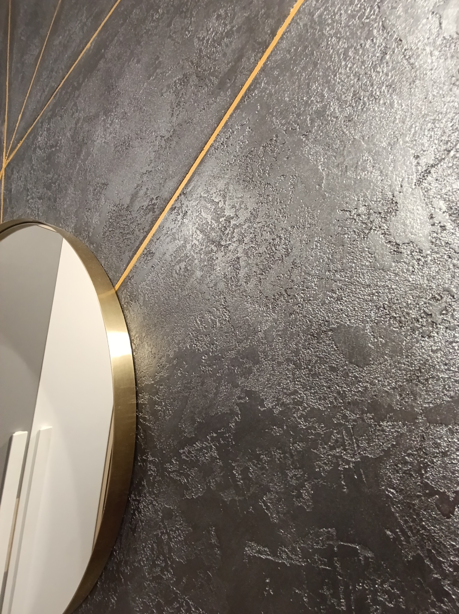 Szara ściana z dekoracyjną fakturą i złotymi liniami, fragment okrągłego lustra z złotą ramą po lewej stronie.