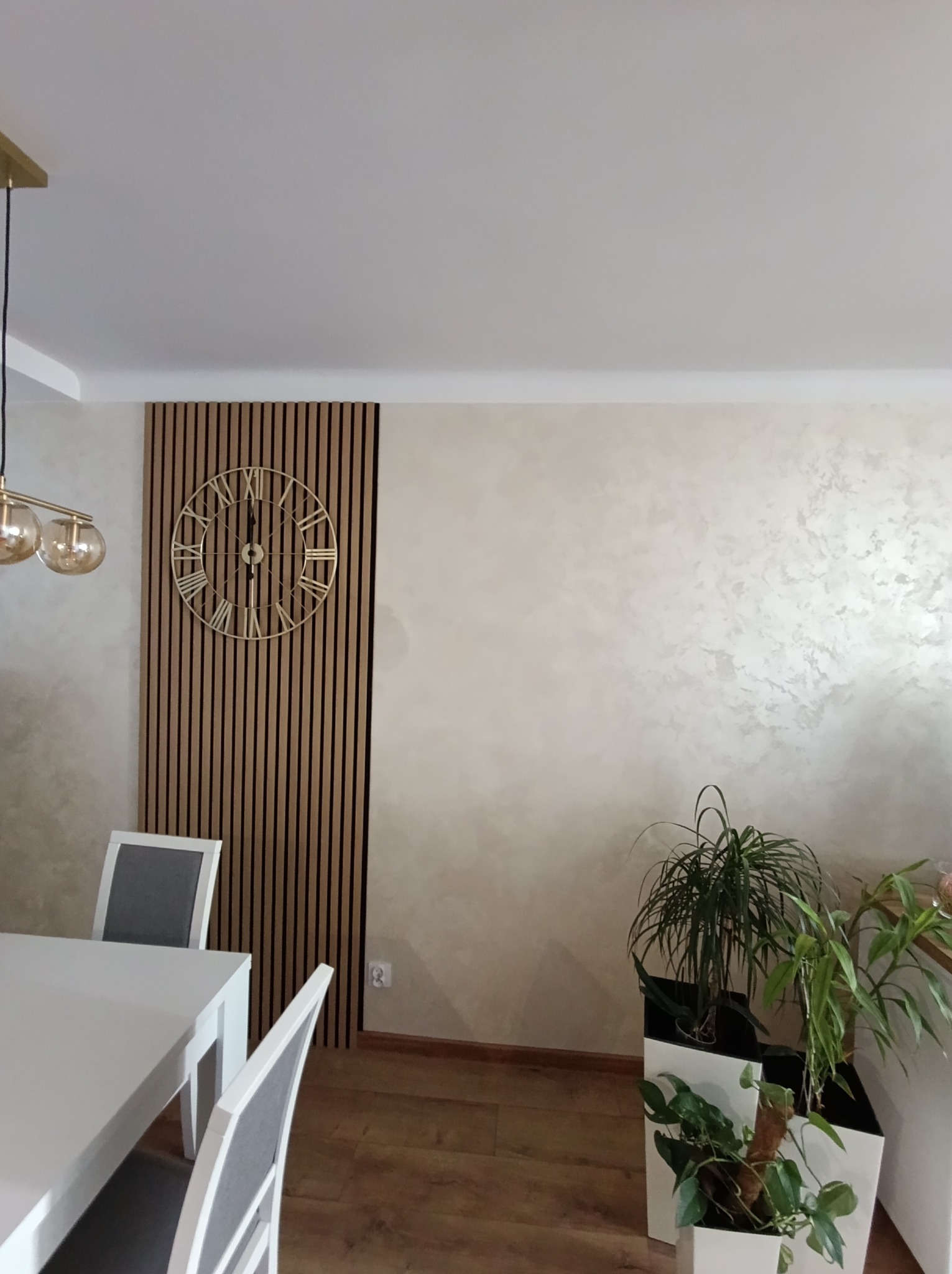 Jadalnia z dekoracyjną ścianą z drewnianych paneli pionowych, na której wisi złoty zegar w stylu retro, obok ściana z efektem dekoracyjnym przypominającym stiuk wenecki, biały stół z szarymi...