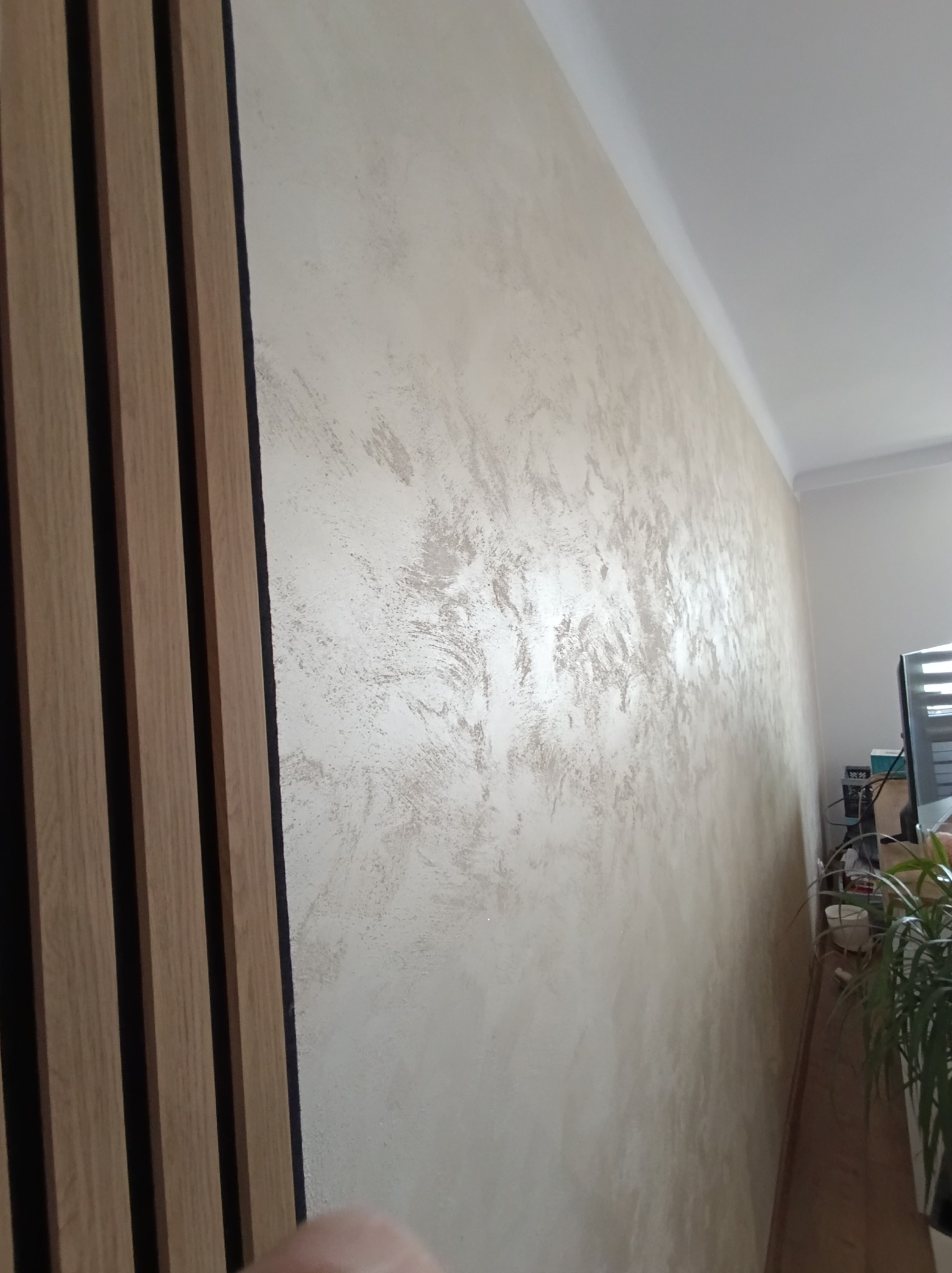 Ściana z dekoracyjną, beżową fakturą i pionowymi panelami drewnianymi z czarnymi wstawkami, widoczne z boku, z fragmentem rośliny w tle.