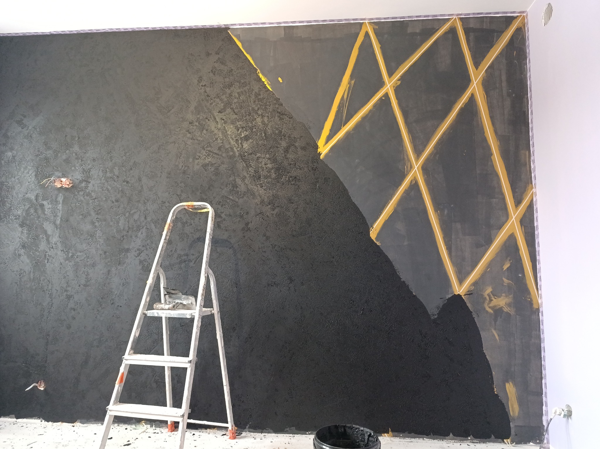 Ściana w trakcie malowania na czarno z geometrycznym wzorem z taśmy malarskiej, widoczna drabina malarska i wiadro z farbą.