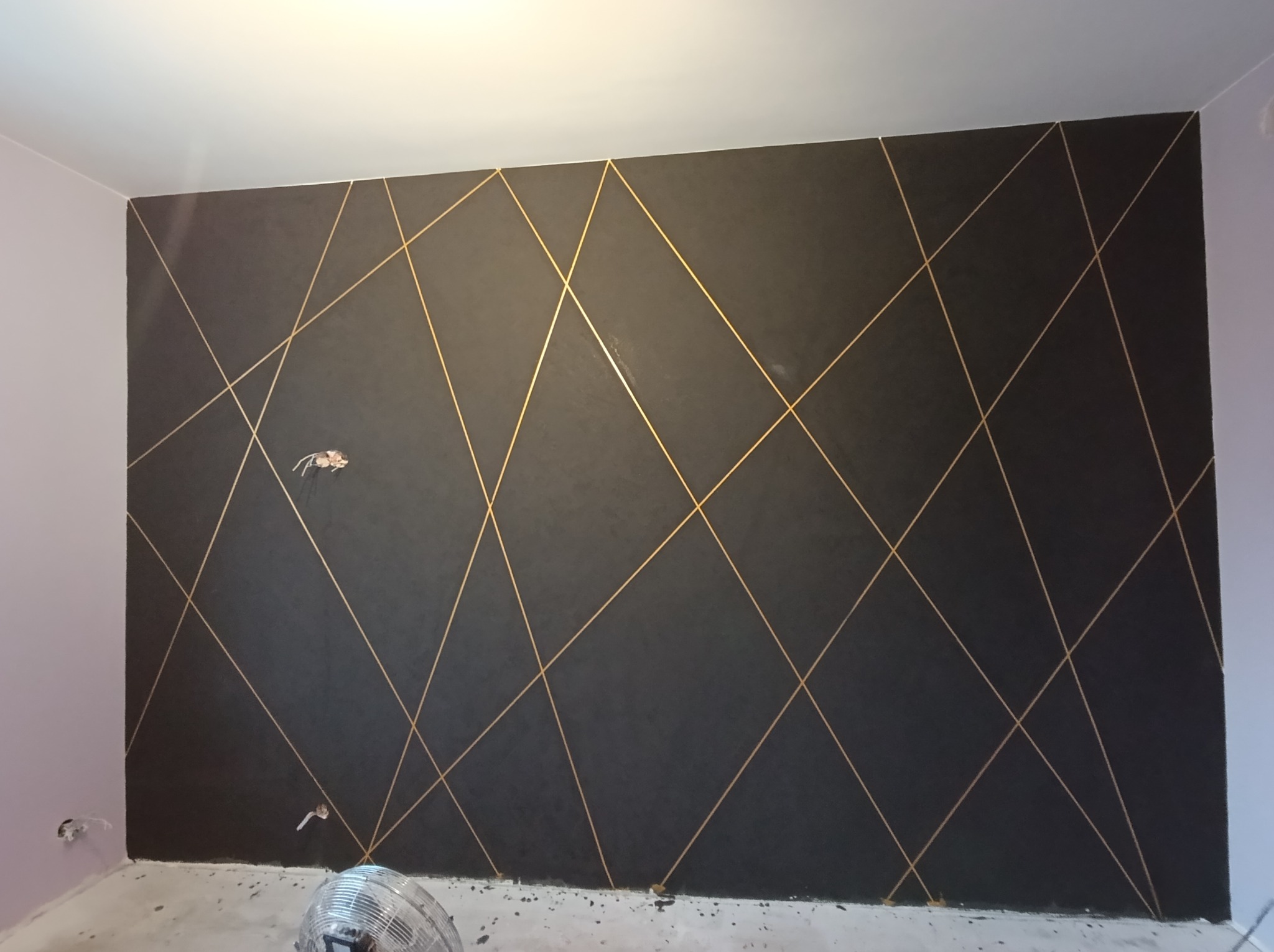 Ciemna ściana z geometrycznym wzorem z cienkich, złotych linii, widoczne niedokończone wykończenie przy podłodze i wystające kable elektryczne.