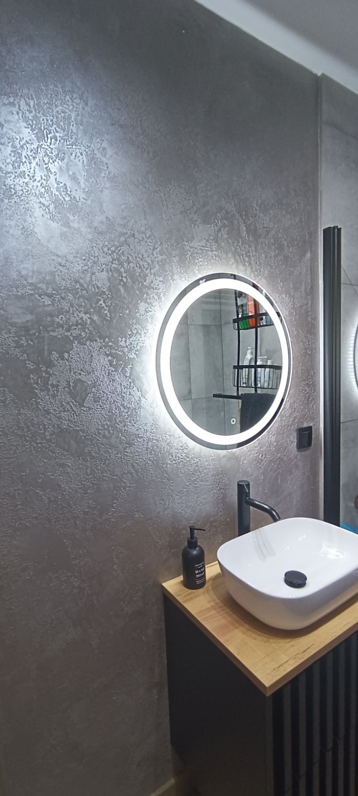 Nowoczesna łazienka w Kutnie z dekoracyjną szarą ścianą o fakturze betonu, okrągłe lustro z podświetleniem LED nad białą umywalką nablatową i czarną armaturą.