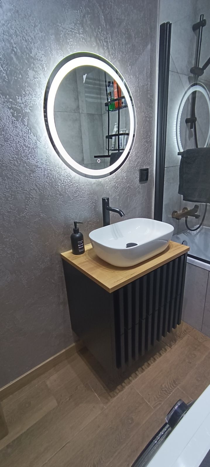 Nowoczesna łazienka z umywalką nablatową na szafce z lamelami, okrągłym lustrem podświetlanym LED i dekoracyjną szarą ścianą z efektem betonu.