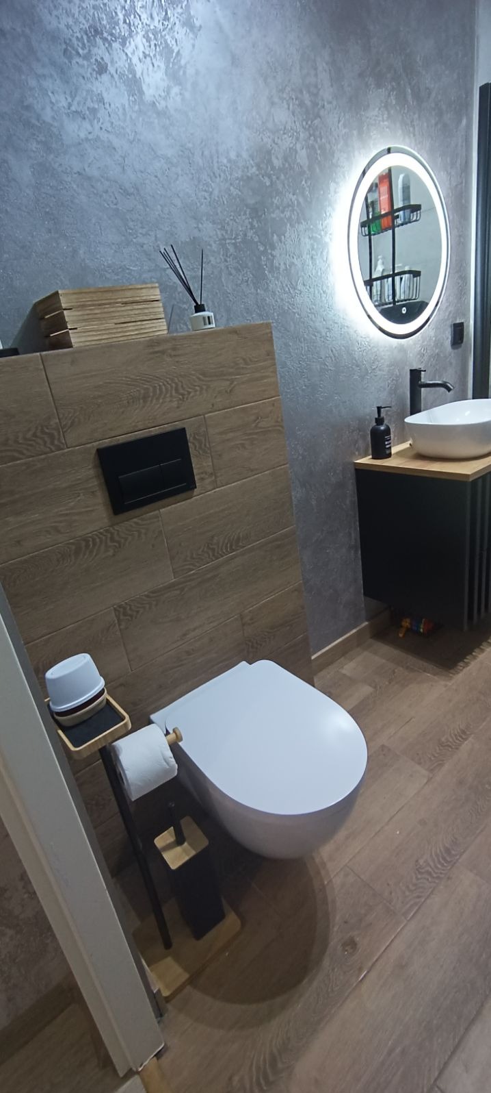 Nowoczesna łazienka z wiszącą toaletą, drewnopodobną okładziną ściany z czarnym panelem sterującym, okrągłym lustrem z podświetleniem LED, umywalką nablatową i szarą ścianą dekoracyjną.
