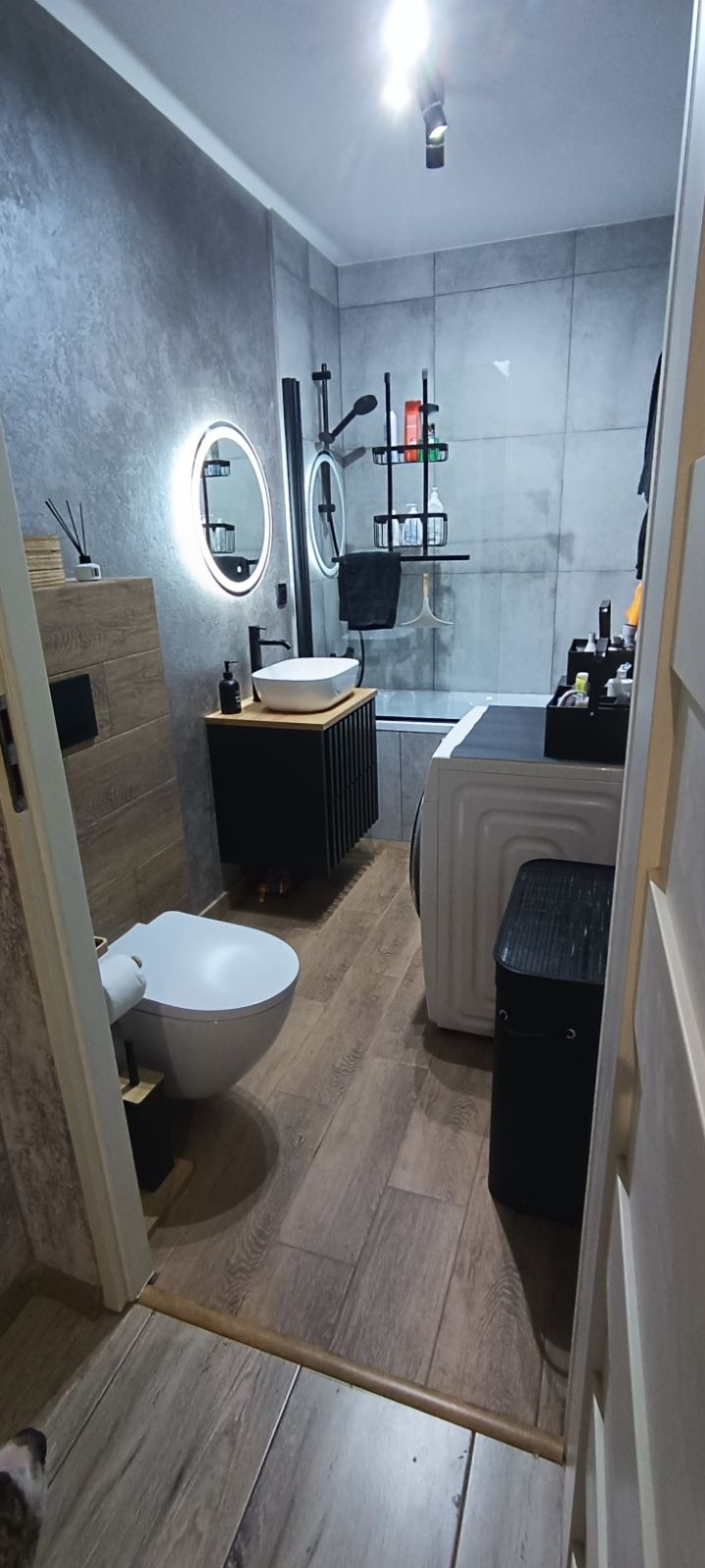 Nowoczesna łazienka z szarymi płytkami imitującymi beton, drewnopodobnymi panelami na podłodze, okrągłym lustrem z podświetleniem LED, umywalką nablatową na czarnej szafce, białą toaletą wiszącą...