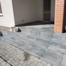 Wykończone schody wejściowe do budynku, wyłożone szarymi płytkami, z widocznym słupem z czerwonej cegły i kostką brukową przed schodami. Dwa wiadra po farbie stoją na górnym stopniu.