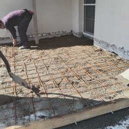 Kozik - Przygotowanie podłoża pod wylewkę betonową: siatka zbrojeniowa ułożona na warstwie betonu, widoczny pracownik w koszuli w kratę i poziomica przy ścianie budynku z oknem.