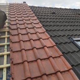 Kozik - Układanie nowych, brązowych dachówek na dachu, widoczny fragment starego, czarnego pokrycia oraz drewniana konstrukcja łatowania.