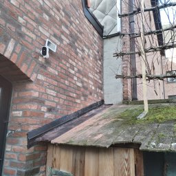 Kozik - Czerwona ceglana ściana budynku z zamontowaną kamerą monitoringu, przylegająca do zadaszonej konstrukcji z mchem na dachu.