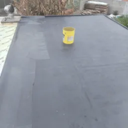 Płaski dach pokryty czarną papą z widocznymi łączeniami i łatami, na środku stoi żółte wiadro, w tle kominek wentylacyjny i zabudowania gospodarcze.