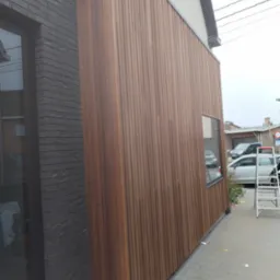 Nowoczesna elewacja budynku z drewnianą okładziną pionową w ciepłym odcieniu brązu, kontrastująca z ciemną, ceglaną ścianą obok okna. Na chodniku widoczna drabina i narzędzia.